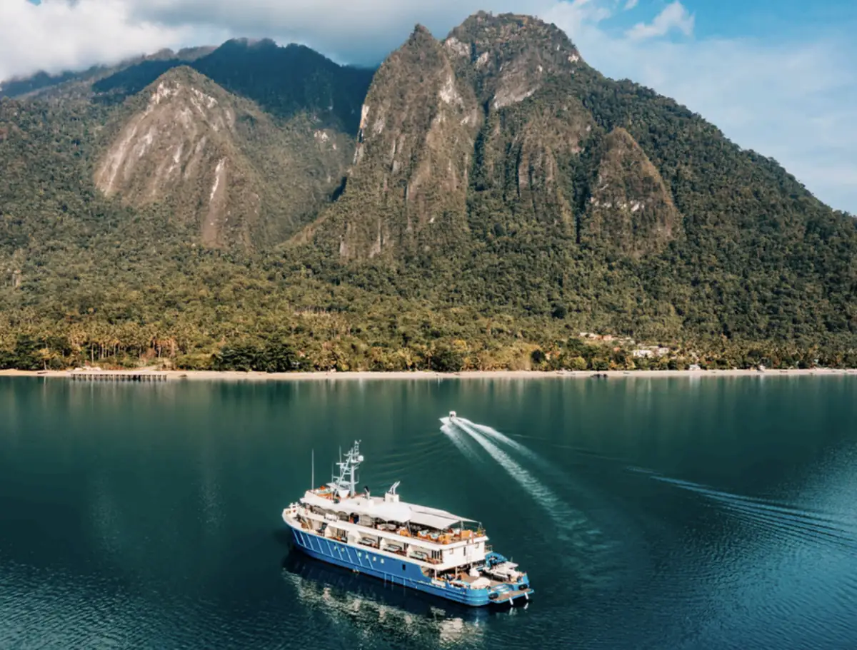 Kudanil Explorer expedition yacht anchored near a lush mountainous coastline in Indonesia.