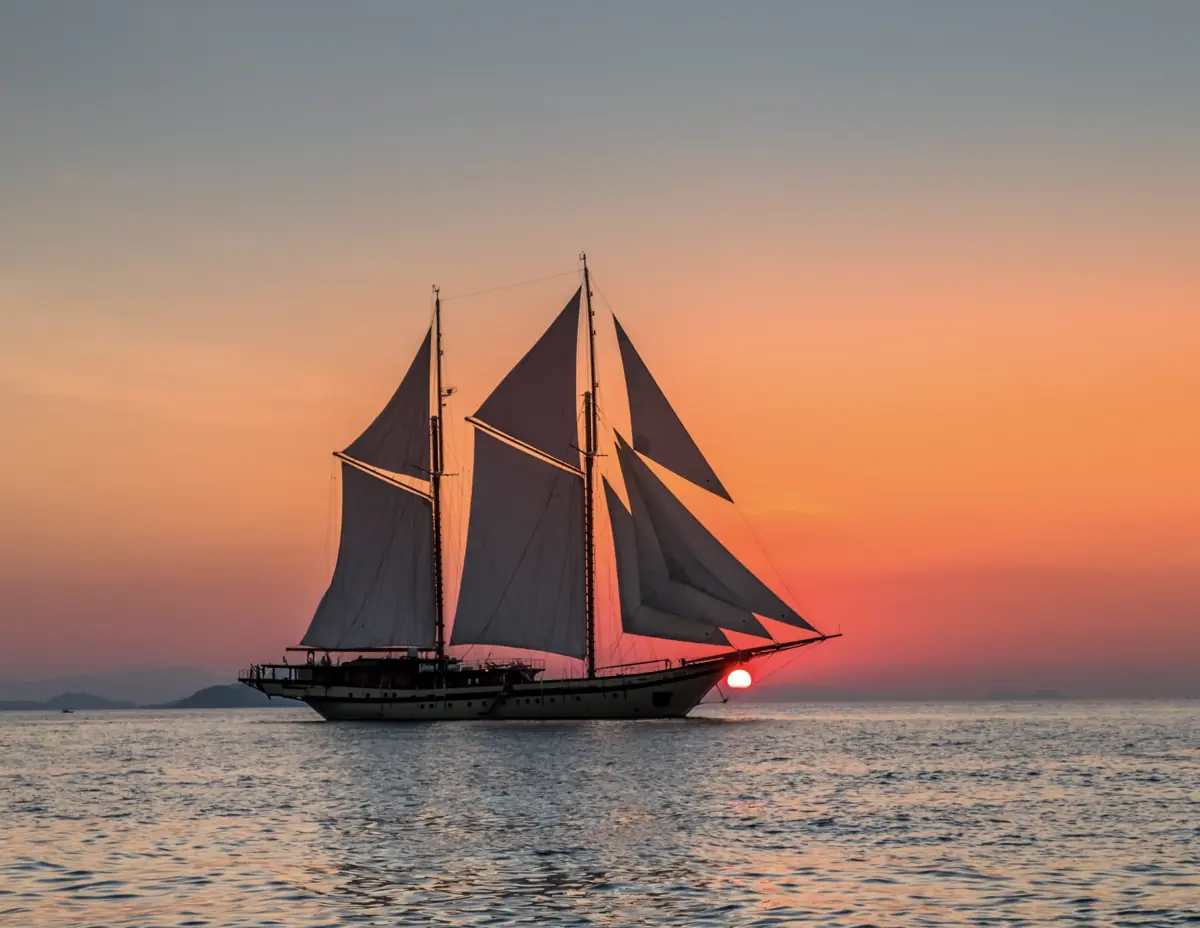 Lamima phinisi yacht sailing at sunset in Komodo National Park with glowing skies.